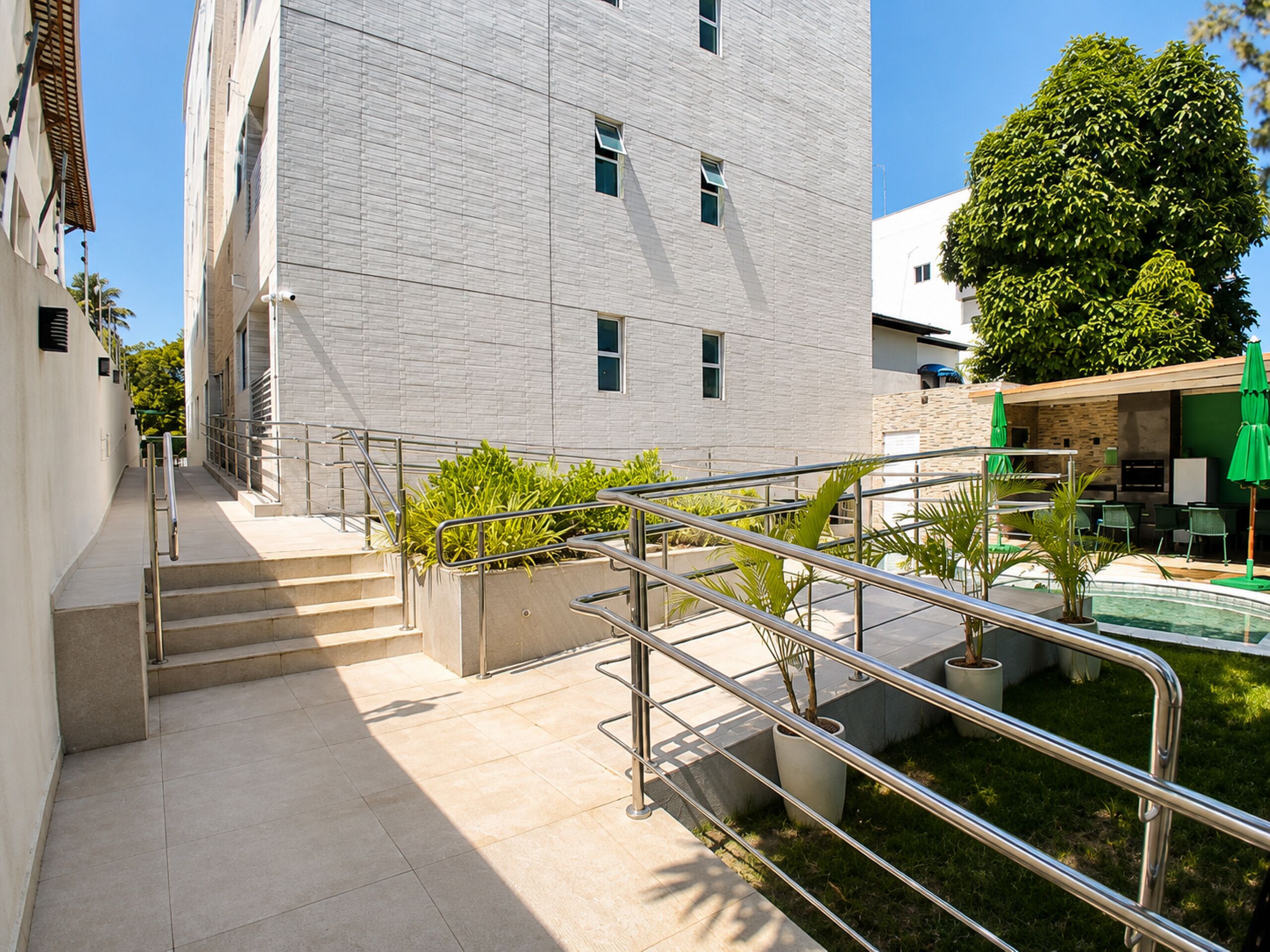 Un edificio moderno con una escalera que conduce a una zona en la azotea, rodeado de vegetación y mobiliario de exterior.