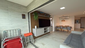 A modern living room with a flat-screen TV, a dining area with a table and chairs, and a kitchen area with a refrigerator.
