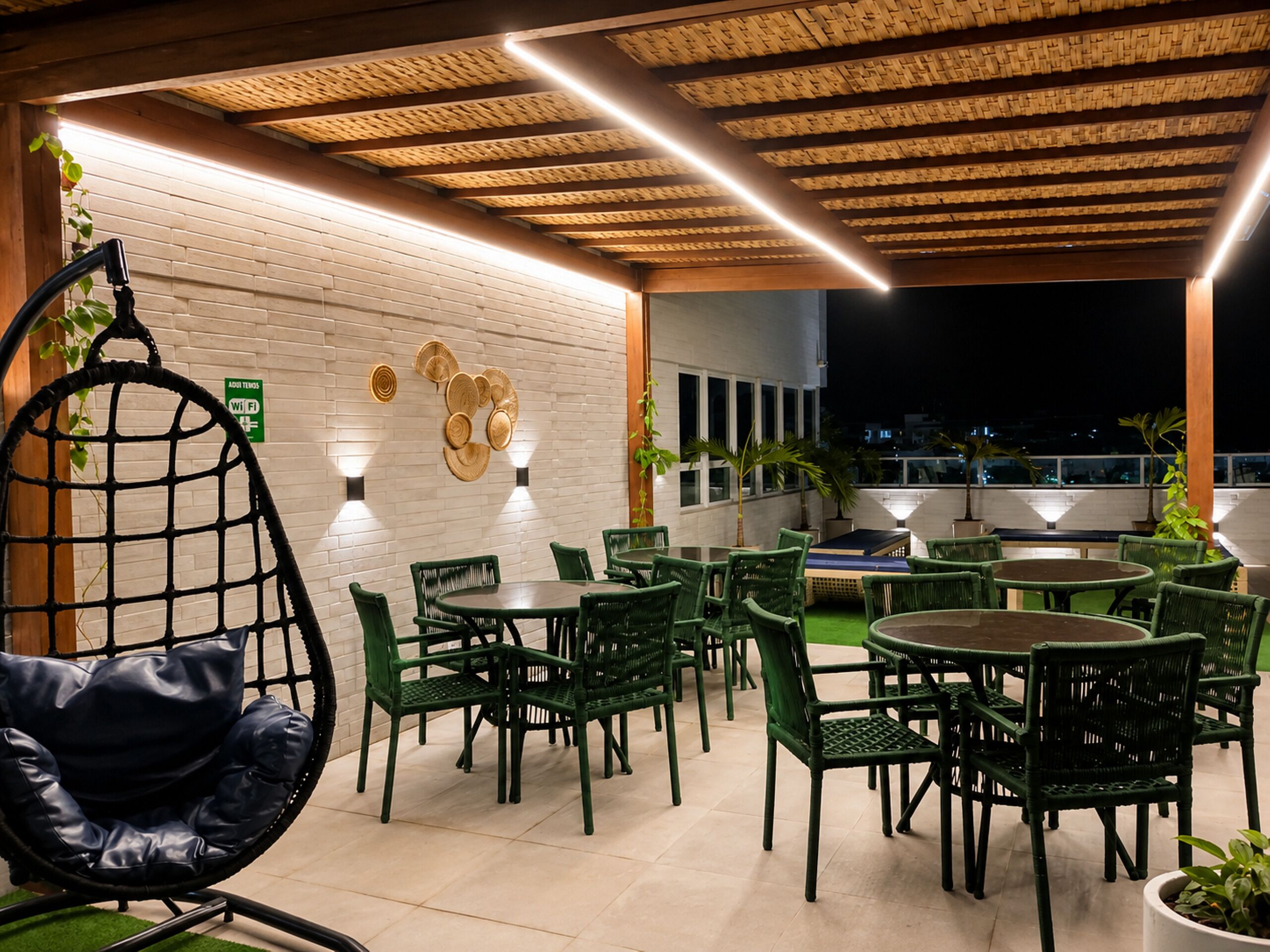 Een terras buiten met groene stoelen en tafels, een hangstoel en een bakstenen muur met decoratieve kunst.