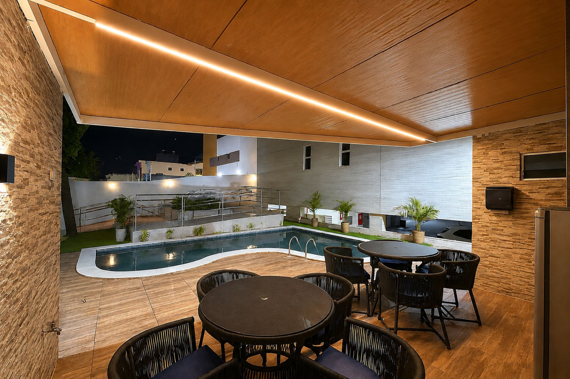 Un espace piscine extérieur doté d’une terrasse en bois, d’une table ronde avec des chaises, et d’une cuisine moderne équipée d’un four encastré.