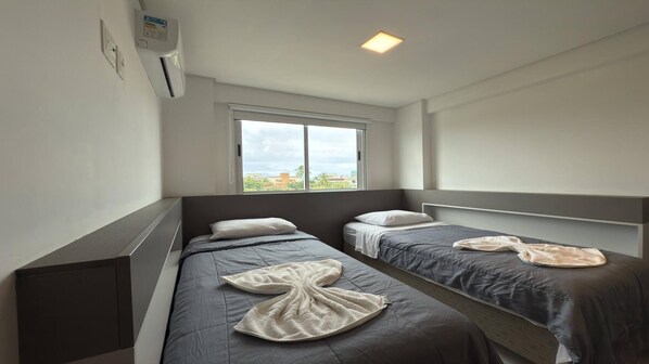 A hotel room with two single beds, a window with a city view, a ceiling light, and a wall-mounted air conditioner.