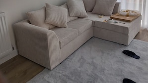 A modern living room with a beige sectional sofa, a white rug, and a large window with sheer curtains. - Room-charming Loughton Apartment With Ba (Loughton)
