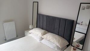 A neatly made bed with white linens, a grey upholstered headboard, and a bedside table with a lamp and a vase. - Room-charming Loughton Apartment With Ba (Loughton)
