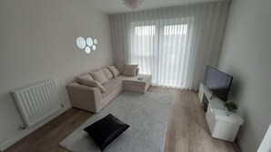 A modern living room with a beige sectional sofa, a black accent pillow, a white coffee table, a flat-screen TV, and a radiator. - Room-charming Loughton Apartment With Ba (Loughton)
