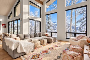 A spacious living room with a large sectional sofa, a patterned rug, and a view of snow-covered trees through large windows.