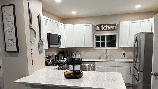 Eine moderne Küche mit weißen Schränken, einer Marmorinsel mit drei Barhocker und Edelstahlgeräten. Ein Schild an der Wand zeigt das Wort 'kitchen'.