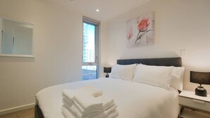 A hotel room with a large bed, a nightstand with a lamp, a window with a view of buildings, and a painting on the wall. - Amyra Residences on Rowlands Place - Unit 705 (Adelaide)