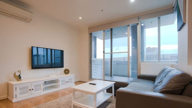 Un salon moderne doté d’un téléviseur à écran plat, d’une console blanche, d’un canapé beige et d’une grande fenêtre offrant une vue sur la ville.
