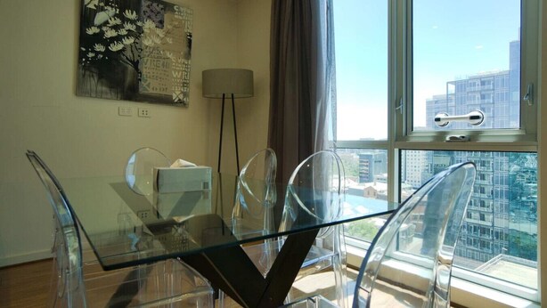 A modern dining area with a glass table and chairs, a painting on the wall, and a view of the city through large windows.