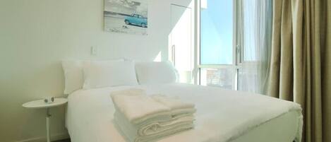 A hotel room with a large bed, a small bedside table, a window with curtains, and a painting on the wall.