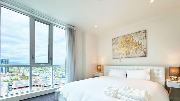 Hotellihuone, jossa on suuri sänky, kaksi yöpöytää valaisimineen, näkymä kaupunkiin suurista ikkunoista sekä seinällä abstrakti taideteos.