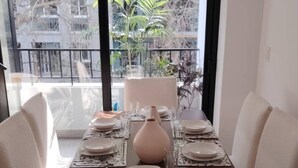 Una mesa de comedor dispuesta con platos, vasos y un florero, ubicada frente a un gran ventanal con vistas a edificios y árboles.
