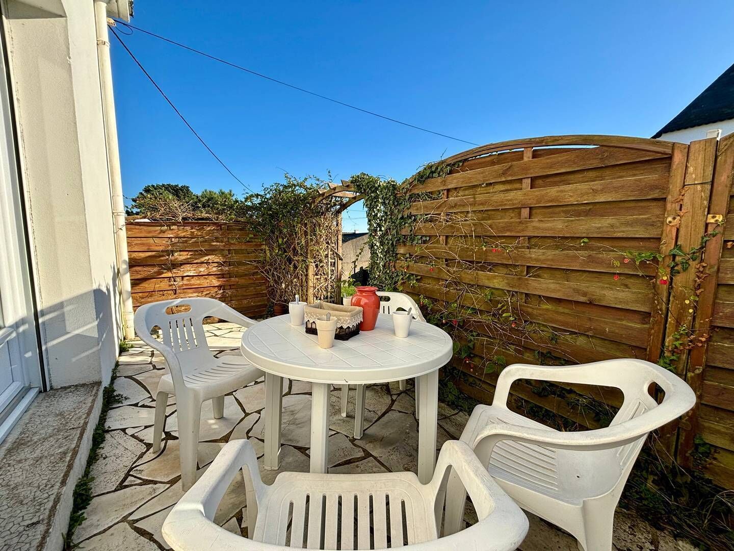 Une terrasse avec une table ronde et quatre chaises en plastique blanc, entourée d’une clôture en bois et de verdure.