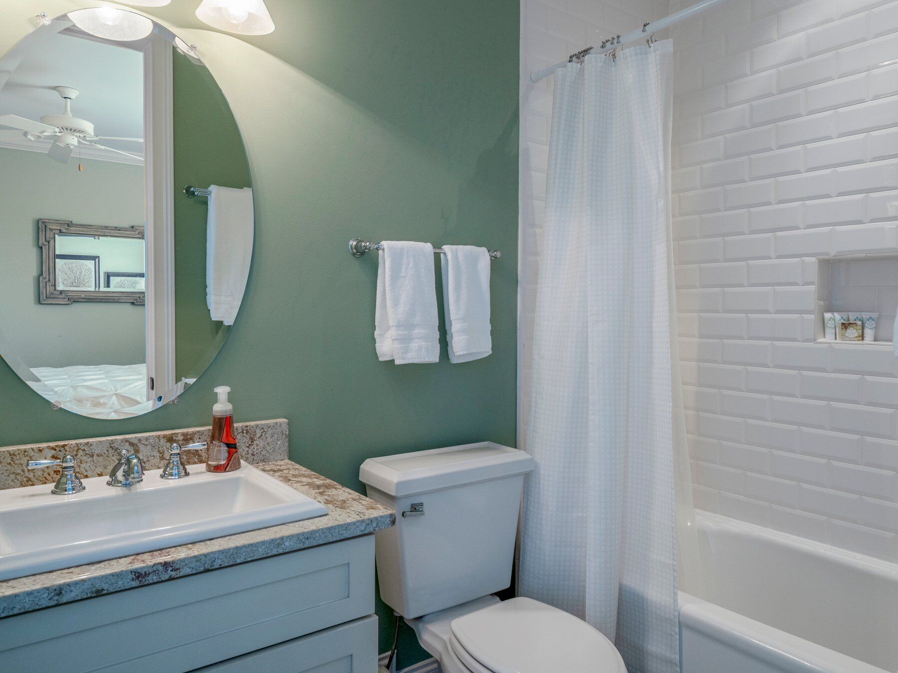Un bagno con il water bianco, un lavandino con piano in marmo, uno specchio rotondo e una doccia con tenda bianca.