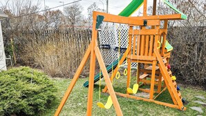Une balançoire en bois avec un toboggan, entourée d’une clôture et de végétation.