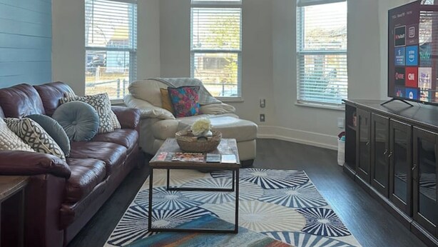 A spacious living room with a leather sofa, a coffee table, and a chandelier. There are three large windows providing natural light. - The Azure Charm — Amazing 3BR 2BA Home 1 Mile to Fort Sam Houston & The Pearl (San Antonio)