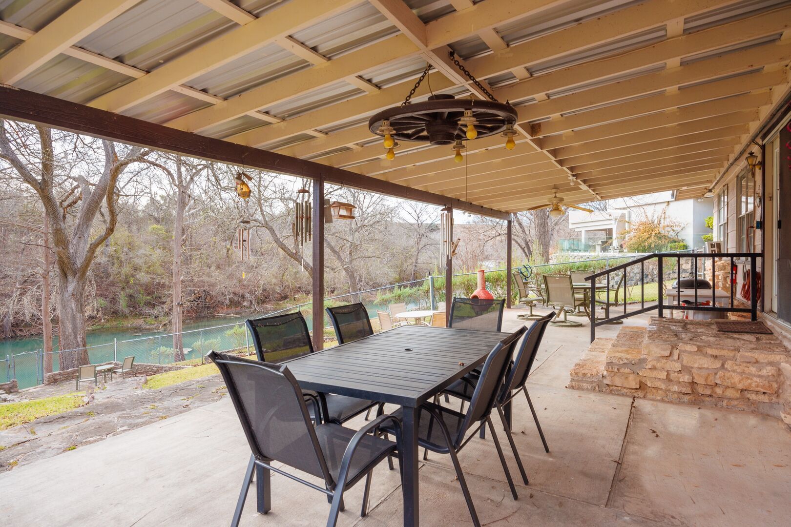Une terrasse extérieure avec une table et des chaises, un espace couvert avec un plafond, et une vue sur une rivière et des arbres.