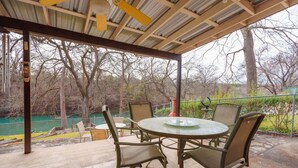 An outdoor patio with a round table and chairs, a metal roof, and a view of trees and a body of water.