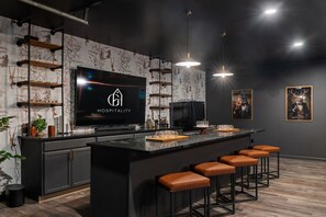 A modern kitchen with a large flat-screen TV, wooden shelves, and a bar area with stools. - The Distillery - Golf Sim, Hot Tub, Game Room (Sugar Grove)