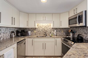 A modern kitchen with white cabinets, a granite countertop, a stainless steel sink, a microwave, a coffee maker, and a kettle. - Sea Rose: Oceanfront Condo, Great Ocean Views, Community Pool (Nags Head)