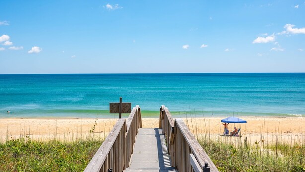 Ein Holzsteg führt zu einem Strand mit blauem Himmel und ruhiger See.