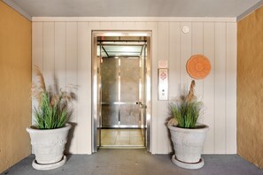 An elevator with a metallic interior, flanked by two large potted plants with green foliage. - Sea Rose: Oceanfront Condo, Great Ocean Views, Community Pool (Nags Head)