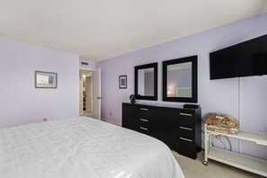 A bedroom with a bed, a black dresser, two mirrors, a television, and a bedside table with a blanket.