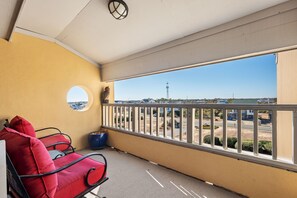A balcony with a yellow wall, a red cushioned chair, a small blue pot, and a view of a road and distant buildings. - Sea Rose: Oceanfront Condo, Great Ocean Views, Community Pool (Nags Head)