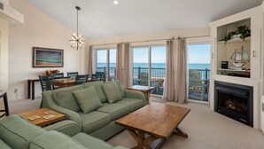 Ein geräumiges Wohnzimmer mit einem grünen Sofa, einem Holztisch und einem Kamin. Es gibt eine Essecke mit Tisch und Stühlen, und große Fenster mit Blick auf das Meer.
