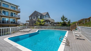 A swimming pool with a wooden deck, lounge chairs, and a stone building in the background. - Sea Rose: Oceanfront Condo, Great Ocean Views, Community Pool (Nags Head)