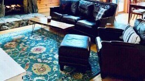Interior acollidor d'una cabana amb una llar de pedra, un sofà de pell negra i una finestra gran.
