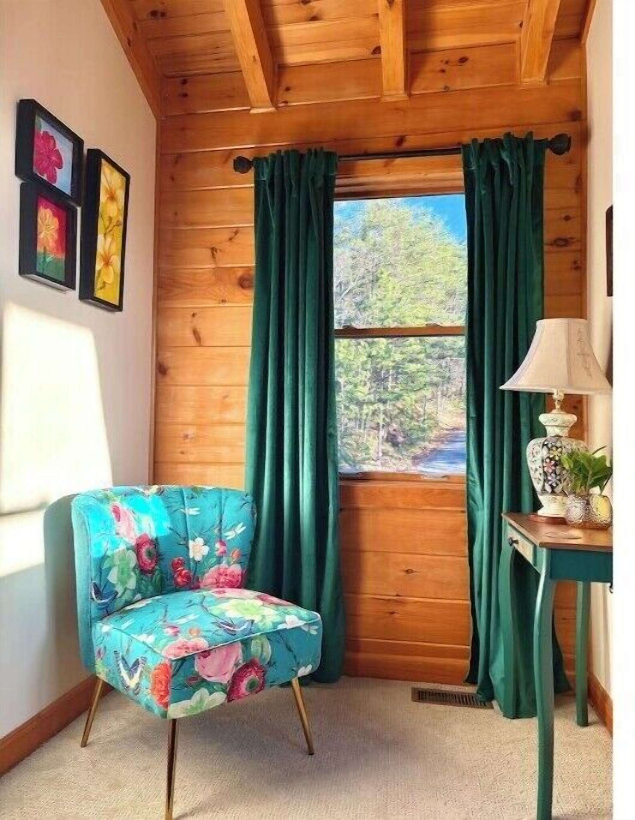 Ein Eckzimmer mit Holzwänden, einem Fenster mit grünen Vorhängen, einem floralen Sessel und einem kleinen Tisch mit einer Lampe.