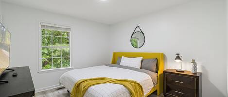 A bedroom with a bed, bedside table, lamp, and a window with a view of greenery.