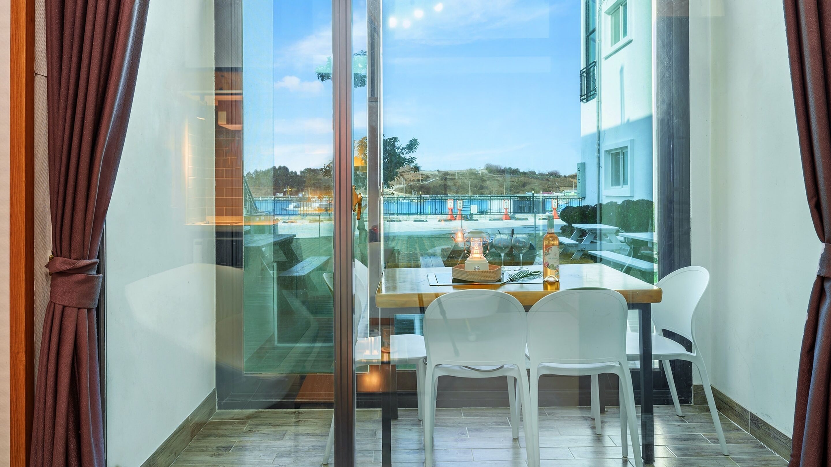 A balcony with a glass door, white plastic chairs, a wooden table, and a bottle of wine.