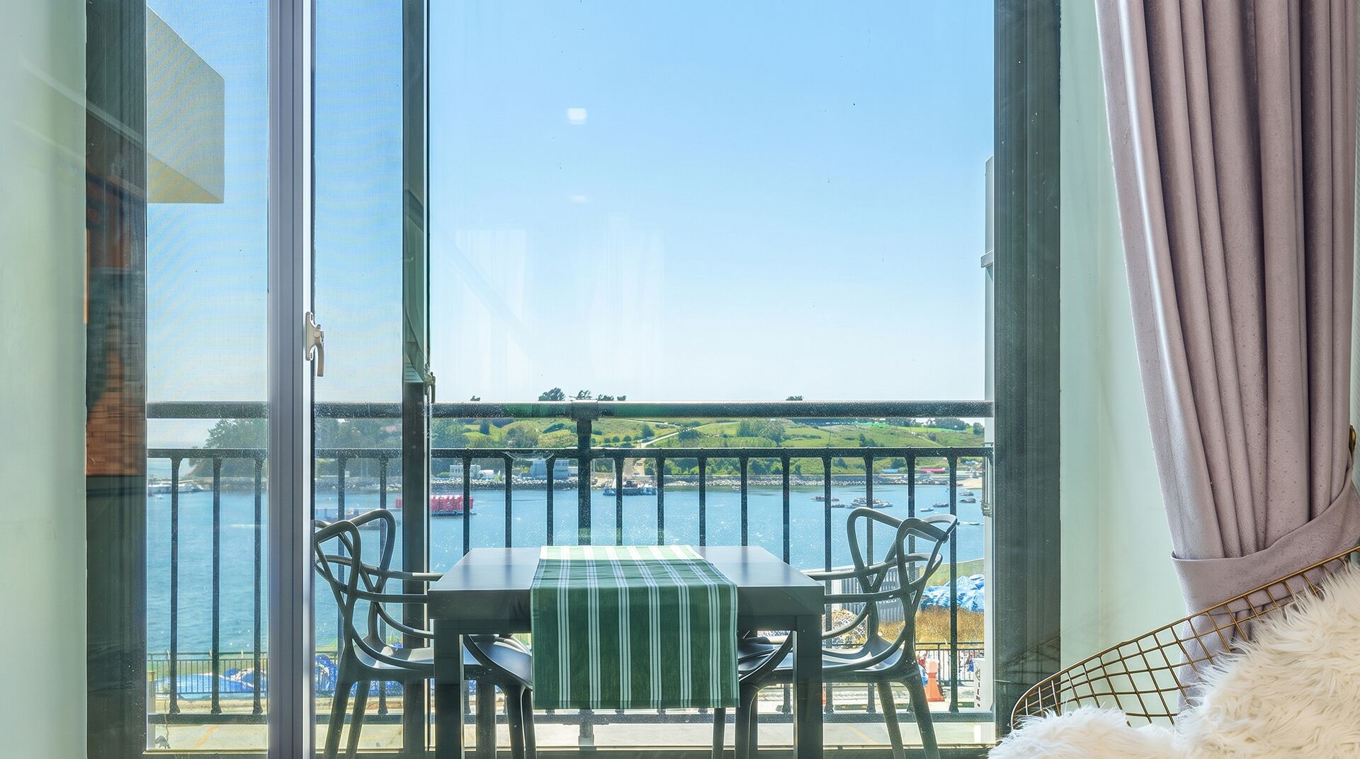 A balcony with a table and chairs, overlooking a body of water with boats and a distant landscape.