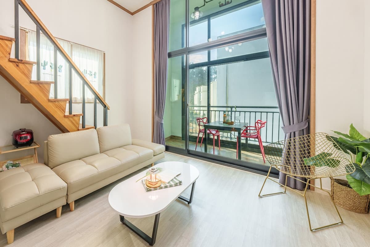 A modern living room with a sofa, coffee table, and a dining area with red chairs. A staircase with wooden railings leads to an upper floor.