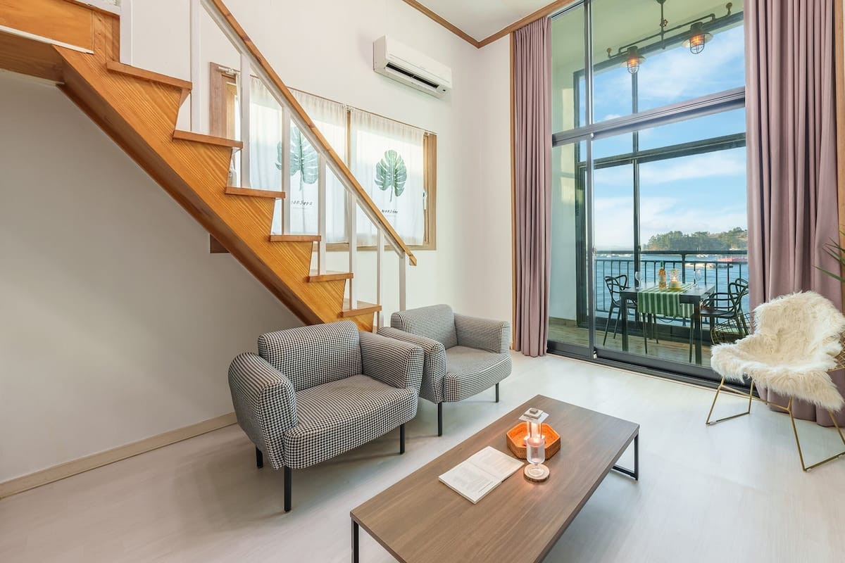 A modern living room with a staircase, large windows, and a view of the sea.