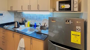 A compact kitchen with light wooden cabinets, a stainless steel refrigerator, a microwave, and a sink with modern fixtures. - Room-simple, Modern & Cozy Condo in Lafa (Iloilo)