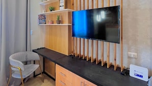 A modern living room with a flat-screen TV mounted on a wooden wall, a grey upholstered chair, wooden cabinets, and a small shelf with decorative items. - Room-simple, Modern & Cozy Condo in Lafa (Iloilo)