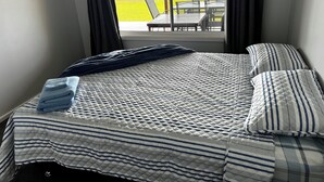 Una cama bien hecha con un edredón a rayas y almohadas, una ventana con cortinas, y vista a un patio exterior con sillas y una mesa.