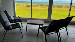 Una habitación con dos sillas y una mesita pequeña, ventanas amplias con vistas a un campo verde y al océano.