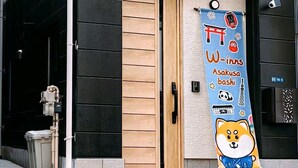 A building with a wooden door, a banner with Japanese text and a Shiba Inu illustration, and a small black sign with numbers.