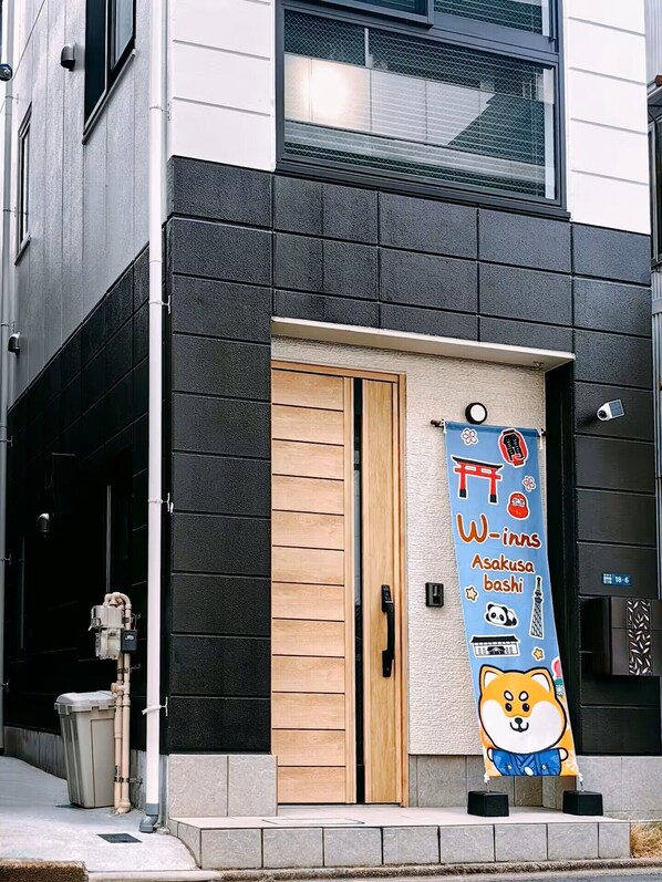 Un edificio con puerta de madera, un cartel con texto japonés e ilustración de Shiba Inu, y una pequeña placa negra con números.