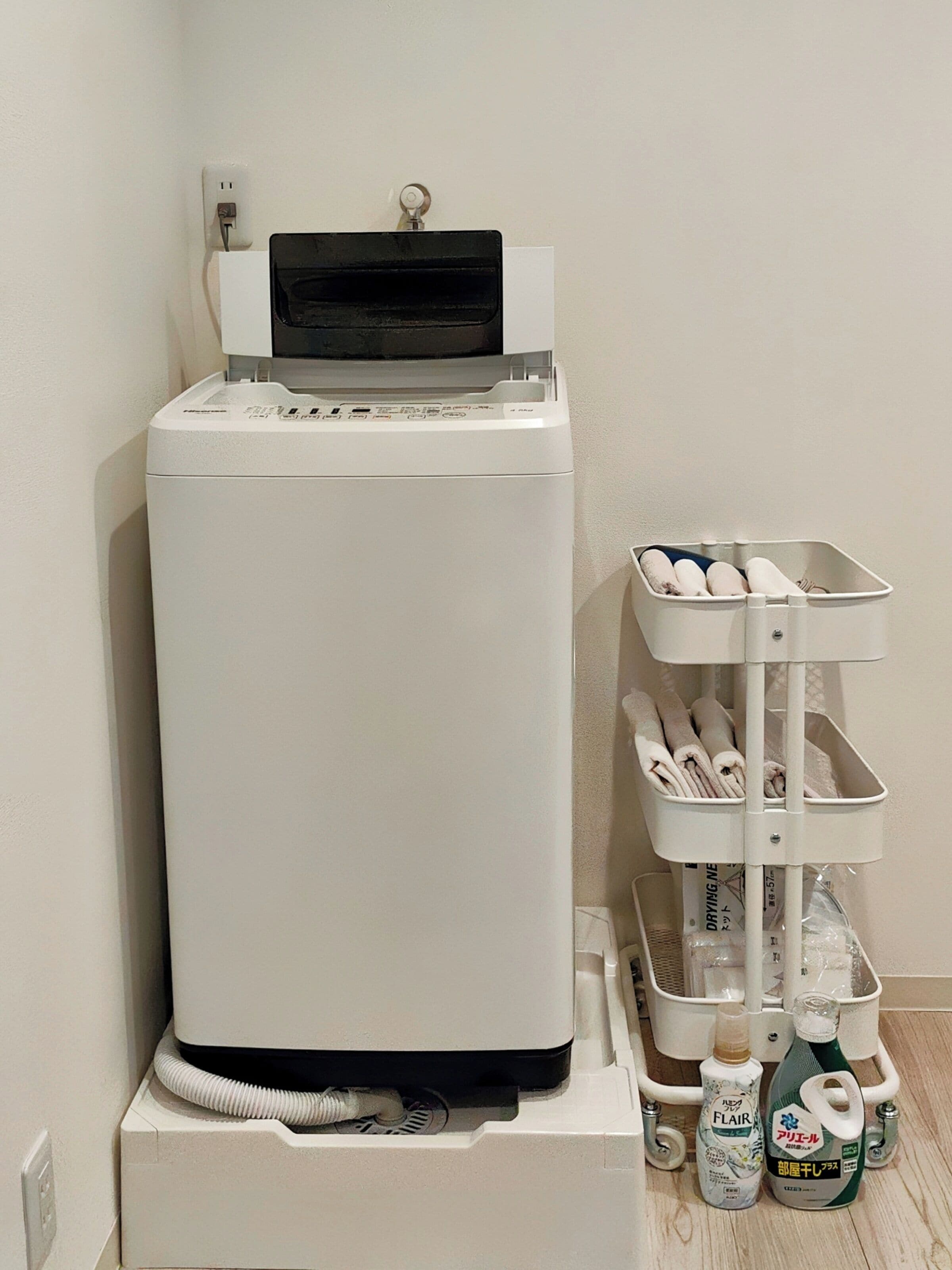 A white refrigerator with a black top, a power outlet on the wall, and a white shelf unit with bottles and towels.