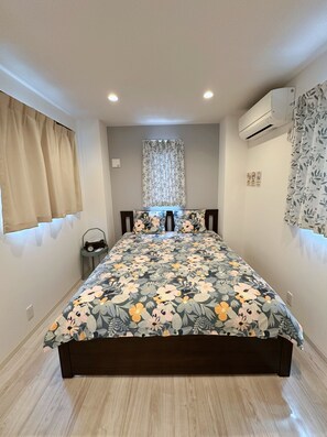 Un dormitorio con edredón estampado de flores, una mesita de noche pequeña con un reloj, una ventana con cortinas color beige y un equipo de aire acondicionado en la pared.