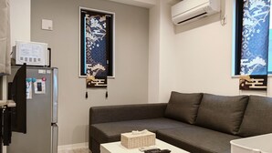 A compact living space with a grey sofa, a small white table, a refrigerator, and an air conditioning unit mounted on the wall.