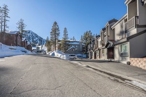 Une rue résidentielle où le sol est recouvert de neige, avec des maisons dotées de balcons et une montagne en arrière-plan.