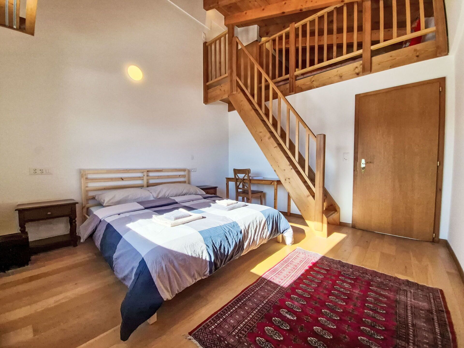 A bedroom with a wooden bed, a small table, a chair, a wooden staircase, and a door.