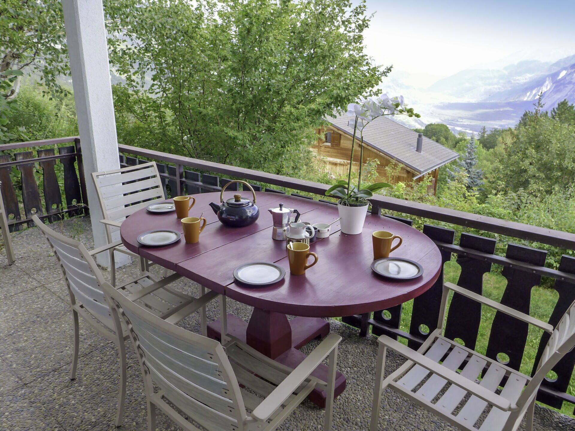 Une table ronde avec des assiettes, des tasses et une théière, placée sur un balcon d’où l’on peut admirer une maison et des montagnes.