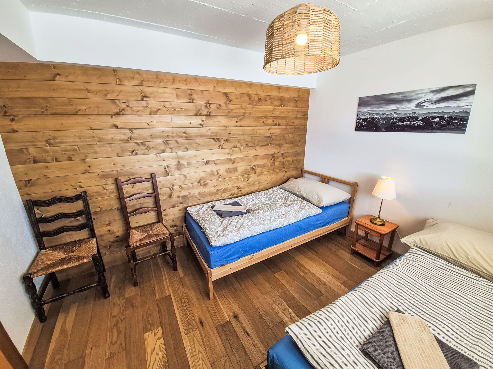 Une petite pièce chaleureuse avec un mur en bois, un lit simple avec un matelas bleu, une chaise, une petite table avec une lampe et un tableau encadré accroché au mur.
